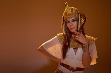 Mısır kıyafetli, başlıklı, çenesine dokunan ve kahverengi arka planda poz veren şık bir kadın.