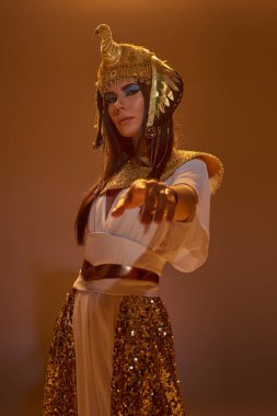 Woman in egyptian attire pointing with finger at camera and standing on brown background with light clipart