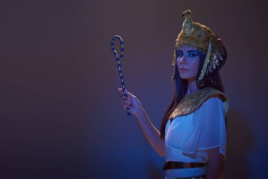 Brunette woman in egyptian attire and headdress holding crook on brown background with blue light clipart
