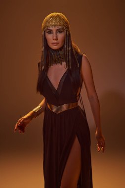 Woman in egyptian look and necklace looking at camera while standing on brown background with light clipart