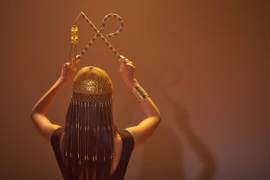 Back view of brunette woman in egyptian headdress holding crook and flail on brown background clipart