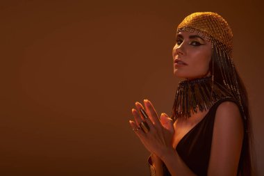 Woman in golden headdress and egyptian look looking at camera isolated on brown with light clipart