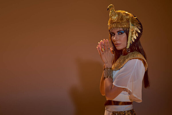 Brunette woman in Egyptian attire looking at camera while posing on brown background with shadow