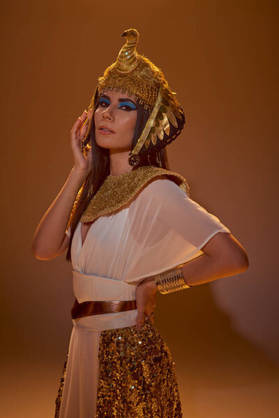 Stylish brunette woman in Egyptian attire and headdress holding hand on hip on brown background