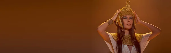 stock image Fashionable woman in Egyptian attire touching headdress in lighting on brown background, banner