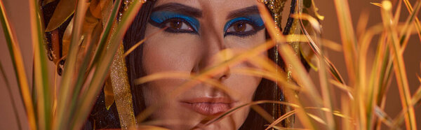 Woman with makeup and egyptian headdress looking at camera behind plants isolated on brown, banner
