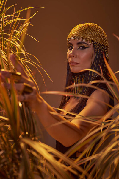Stylish brunette woman in egyptian look touching plants and looking at camera isolated on brown