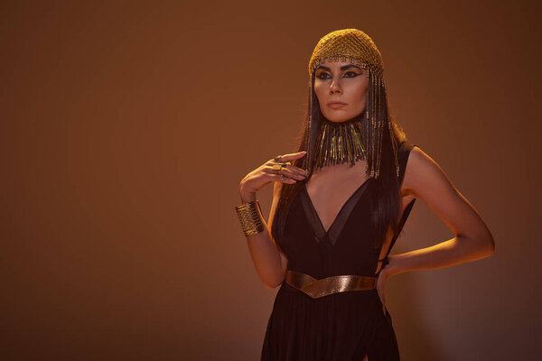 Trendy woman in egyptian look and necklace holding hand on hip while standing on brown background