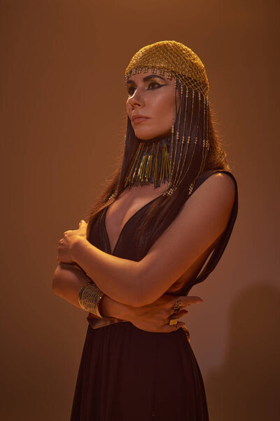 Trendy woman in egyptian look and headdress posing and looking away on brown background