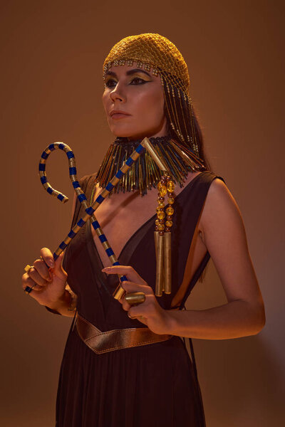 Elegant woman in egyptian golden headdress and look holding crook and flail on brown background