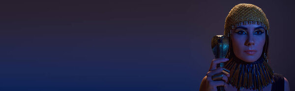 Woman with makeup and egyptian headdress holding crook in snake shape isolated on purple, banner