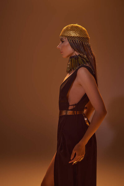 Side view of attractive woman in egyptian headdress and look posing and standing on brown background