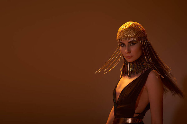 Woman with egyptian makeup and attire looking at camera while posing on brown background with light