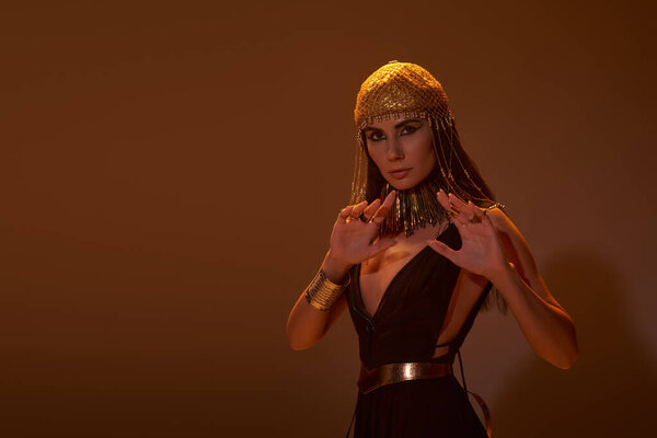 Woman in egyptian dress and headdress posing and looking at camera on brown background with light