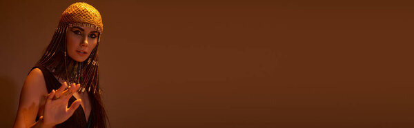 Stylish woman in egyptian costume and headdress posing on brown background, banner