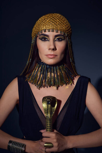 Portrait of woman in egyptian look and headdress holding crook in snake shape on blue background
