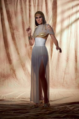 Trendy model in egyptian attire and pearl top posing while standing on abstract background clipart