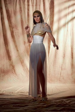 Trendy model in egyptian attire and pearl top posing while standing on abstract background clipart