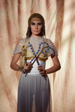 Mısırlı kostümlü ve incili bluzlu bir kadın, soyut arka planda poz verirken elinde dolandırıcı ve kırbaç tutuyor.