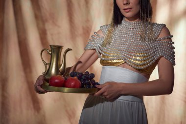 Cropped view of woman in egyptian look holding jug and fruits while posing on abstract background clipart