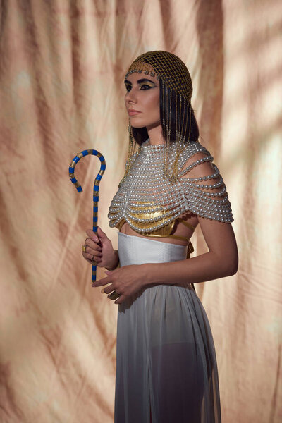 Stylish woman in egyptian look and pearl top holding crook and standing on abstract background