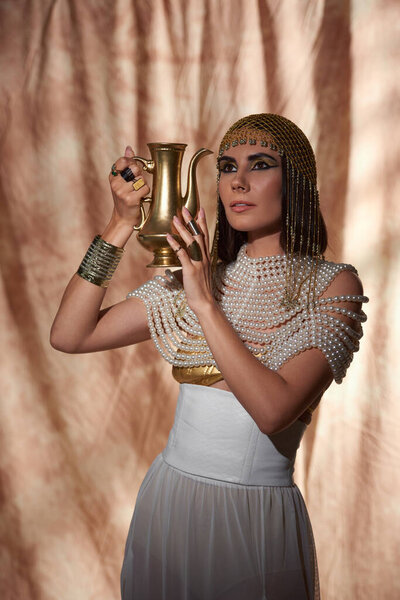 Woman in egyptian look and pearl top holding jug while posing on abstract background