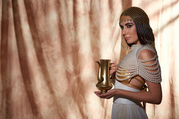 Stylish woman in Egyptian attire and pearl top holding golden jug on abstract background