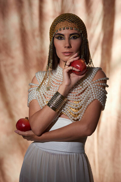 Elegant woman in egyptian attire and headdress holding pomegranates on abstract background