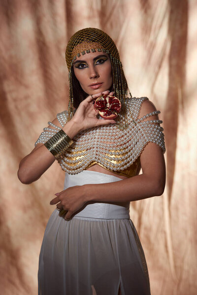 Stylish woman in egyptian attire and pearl top holding cut pomegranate on abstract background