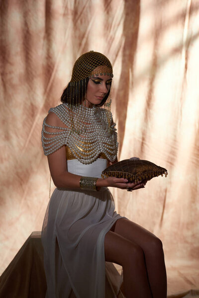 Woman in egyptian look holding pillow with jewelry and gemstones while posing on abstract background