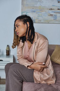 Afro-Amerikan kadın yatakta oturuyor, kendini kötü hissediyor, kameraya, strese, modern yatak odasına bakıyor.