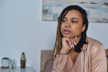 Üzgün Afro-Amerikan kadın yatakta oturuyor, kendini kötü hissediyor, bir bardak su tutuyor, modern yatak odası.