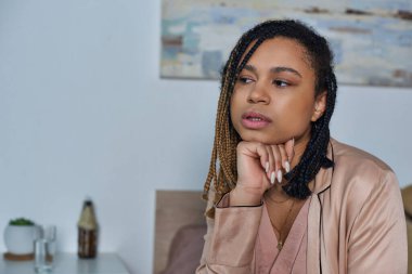 sad african american woman sitting on bed, feeling unwell, holding glass of water, modern bedroom clipart