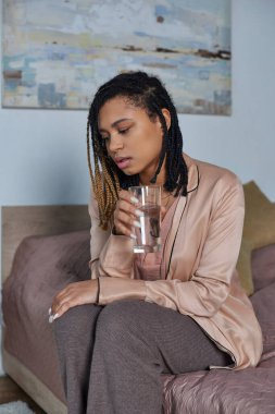 cropped, sad african american woman sitting on bed, feeling unwell, holding glass of water,  bedroom clipart