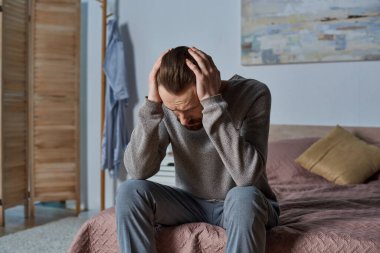 stressed man looking down and sitting on bed in modern bedroom, painting on wall, solitude, concern clipart