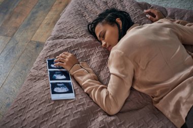 top view of worried african american woman lying on bed near ultrasound, unborn baby, abortion clipart