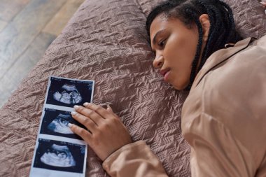 top view of worried african american woman lying on bed near ultrasound,  abortion concept clipart