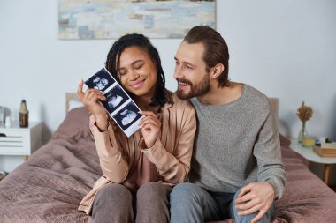 happy interracial husband and wife, future parents, pregnant woman holding ultrasound scan clipart