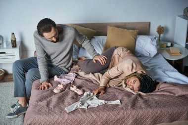 grief, miscarriage concept, man calming african american wife crying near baby clothes, lying on bed clipart