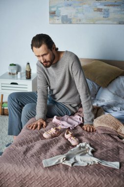 Keder, depresyondaki adam bebek kıyafetlerinin yanında ağlıyor, yatakta oturuyor, düşük konsepti, kalp kırıklığı