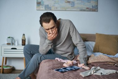 disbelieve, depressed man sitting on bed near baby clothes and ultrasound scan, miscarriage concept clipart