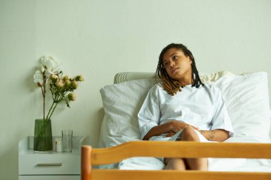 sad african american woman lying in private ward, flowers, glass of water, hospital, miscarriage clipart