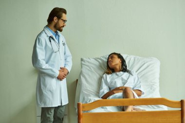 Sakallı doktor, hastane önlüklü, özel koğuşlu, hasta, Afrikalı Amerikalı kadının yanında duruyor.
