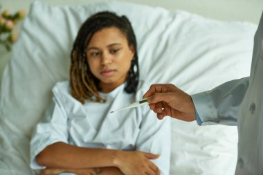 Beyaz önlüklü doktor, termometreyi Afrikalı Amerikalı bir kadının yanında tutuyor. Özel koğuş, hastane.