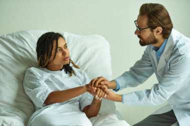 Üzgün Afrikalı Amerikalı kadının elini tutan doktor. Rahatlatıcı hasta, hastanede özel koğuş.