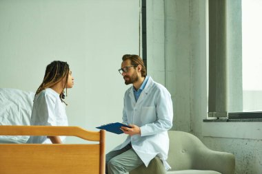 Gözlüklü ve beyaz önlüklü bir doktor elinde pano, Afrikalı Amerikalı bir kadınla konuşuyor, özel koğuş.