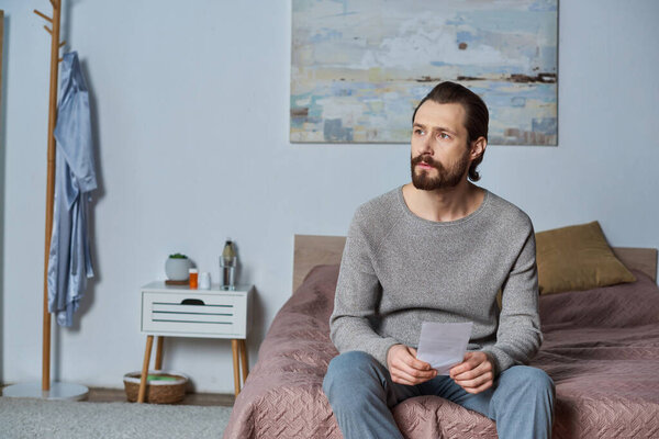 stressed man holding ultrasound photo, sitting on bed, pregnancy, responsibility, making decision