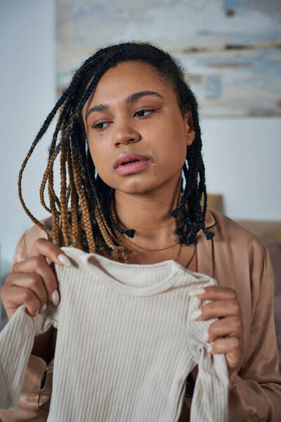 depressed african american woman holding baby clothes and crying in bedroom, miscarriage concept
