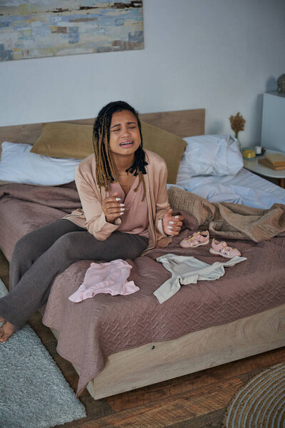 despair, depressed african american woman crying near baby clothes on bed, miscarriage concept