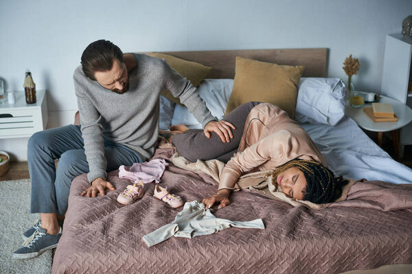 grief, miscarriage concept, man calming african american wife crying near baby clothes, lying on bed